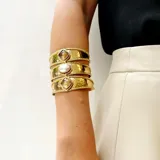 Brazalete ancho de bronce dorado, con textura martillada, compuesto por tres bandas unidas y adornado con tres piedras engarzadas: una central ovalada de nácar blanco y dos laterales cuadradas de nácar blanco.