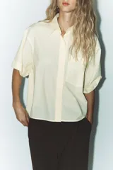 Camisa color crema de cuello solapa y manga corta con vuelta. Tiene un bolsillo de parche en el delantero, bajo asimétrico con aberturas laterales y cierre frontal con botones.
