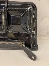 Billetera tipo clutch de cuero charol negro con cierre de cremallera y solapa con tres apliques piramidales.