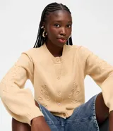 Cardigan de punto color beige, con cuello redondo, cierre frontal de botones, mangas largas con puños acanalados y bordado floral en las mangas y el frente.