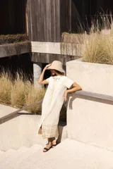 Vestido largo de lino color crudo, con cuello camisero, botones frontales y bordados en contraste que recorren la silueta, incluyendo un detalle estampado en el ruedo.