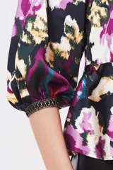Camisa estampada con flores en tonos morados, azules, amarillos y verdes sobre fondo negro. Tiene cuello redondo, mangas abullonadas 3/4 y lazo en la cintura.