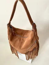 Bolso tipo hobo color tostado, hecho de cuero con flecos largos en el frente y dos bolsillos con cierre. Tiene una manija regulable con hebillas metálicas.