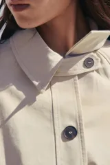 Trench corto color crema con cuello solapa, manga larga con trabilla, bolsillos de doble vivo, forro interior y cinturón lazada en el mismo tejido.