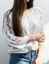 Blusa blanca de rayón con escote redondo, apertura central con botones y mangas largas con bordados florales en tonos azules y naranjas.