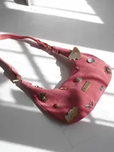 Cartera baguette de gamuza rosa con asa corta y múltiples apliques decorativos: botones, monedas, dijes de metal con forma de sol y caballo, y placa metálica con la marca "Agnes Lenoble".