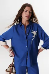 Camisa verde militar de manga larga con puños color crema, cuello clásico y cierre frontal con botones. Tiene un bolsillo en el pecho con bordado de una palmera color crema.