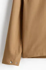 Campera color marrón claro de tejido suave con cuello clásico, cierre frontal con cremallera, bolsillos laterales insertados al bies y puños ajustables con botón a presión.