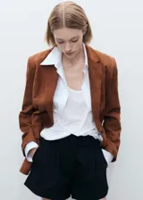 Chaqueta marrón de piel caprina con cuello de pico con solapa, diseño recto, cierre delantero con botones, forro interior, manga larga y dos bolsillos de solapa en la parte frontal.