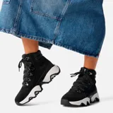 Bota de caña alta Sorel Kinetic Impact, color negro, con cordones y ojales metálicos. Suela de goma blanca con diseño geométrico.