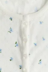 Top de manga corta, corte entallado, en punto pointelle blanco con estampado floral pequeño en tonos azules. Presenta cuello redondo ribeteado y tapeta de botones en el centro del escote.