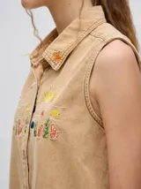 Camisa sin mangas de denim liviano color camel, con cuello camisero y botones frontales. Presenta bordados temáticos marinos en el pecho, incluyendo un sol, olas, una gaviota y vida marina.