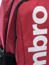 Mochila de color rojo oscuro con base y detalles en negro. Presenta el logo de Umbro en grande en vertical en el frente y el logo pequeño en el panel superior derecho. Incluye bolsillo lateral de malla.
