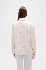 Campera de algodón color crudo, estilo camisa o chaqueta ligera, con bordados en forma de paisley en tono beige claro. Posee cuello camisero, cierre frontal con botones marrones y bolsillos delanteros.