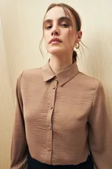 Camisa blanca de manga larga con textura sutil y caída fluida. Presenta cuello clásico, botones forrados a tono y puños con terminación fruncida.