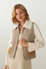 Chaleco corto color beige, de corte recto y abierto, con cuello de piel sintética blanca y apliques de cuero marrón en los bolsillos.