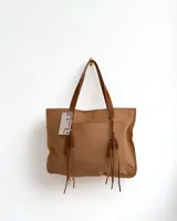 Bolso tipo tote de cuero color marrón claro con textura perforada. Tiene doble asa de cuero marrón y flecos decorativos en los laterales.