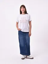 Remera blanca de algodón con cuello redondo y manga corta. Presenta un pequeño bordado en el pecho izquierdo con una figura que parece ser una persona acostada en una tabla de surf o flotador.