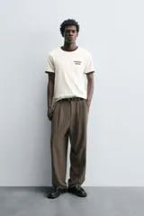 Pantalón negro de corte ancho con cintura elástica, pliegues frontales, bolsillos laterales y traseros de vivo. Tiene detalle de hebilla metálica en la trabilla y cierre frontal con cremallera y botón.