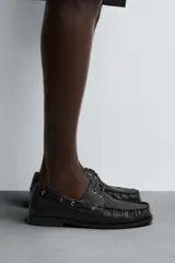 Zapato náutico de vestir color negro, con parte superior lisa, bordón, detalle de ojales metálicos, horma redonda y suela a tono con ligero tacón.