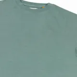 Remera de manga corta color verde azulado, con cuello redondo y corte holgado. Presenta una pequeña etiqueta blanca cosida en el ruedo inferior.
