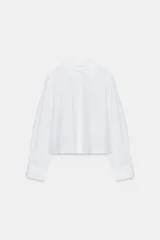 Camisa blanca de popelina con cuello solapa, manga larga abullonada con puño y bajo recto. Cierre frontal con botones ocultos por solapa.