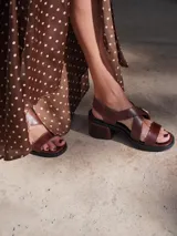 Sandalias bajas de cuero color marrón con tiras anchas cruzadas sobre el empeine y hebilla metálica dorada en forma de estribo.