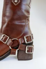 Bota de cuero marrón con caña alta, taco de madera y arnés con hebillas metálicas.