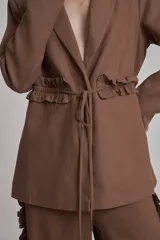 Blazer marrón con solapas de muesca, cierre frontal con lazo y mangas largas. Presenta volados a lo largo de las costuras laterales del pantalón a juego.