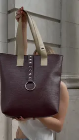 Bolso tote color borgoña con textura símil cuero de cocodrilo y asas color beige. Tiene un aro metálico decorativo, cierre superior y un bolsillo interno.