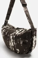 Bolso de hombro tipo bandolera, confeccionado en tejido de algodón con estampado abstracto en tonos blanco y negro. Cuenta con un compartimento principal con cierre de cremallera, un bolsillo interior con cremallera y asa de hombro ajustable.
