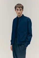 Campera bomber azul marino con cuello elevado acanalado y cierre frontal. Tiene bolsillos laterales y un bolsillo interior, todos con cierre. Los puños y el bajo son acanalados. Está forrada.