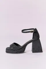 Sandalias de cuero negro con plataforma y tacón cuadrado alto. Presentan una tira delgada sobre los dedos y pulsera tobillera con hebilla dorada.