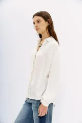 Camisa blanca de manga larga confeccionada en viscosa con acabado sandwash, de textura suave y aterciopelada. Cuello camisero con botón en forma de corazón y frunces en el canesú delantero y la espalda.