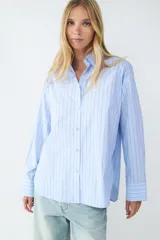 Camisa celeste con finas rayas verticales negras, de corte amplio, manga larga, cuello clásico, cierre frontal con botones y puños abotonados.
