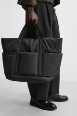 Bolso tipo shopper negro fabricado en tejido técnico. Cuenta con un compartimento principal con cierre de cremallera y un bolsillo interior con cierre. En la parte frontal tiene tres bolsillos. Tiene dos asas de mano.