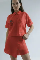 Camisa corta de manga corta color rojo, con doble cartera diagonal con botones de madera, cuello clásico y botamangas marcados. Tiene un bolsillo con aplique de crochet hecho a mano con forma de frutilla.