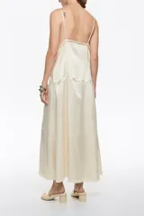 Vestido midi color crema o blanco roto, con tirantes finos y escote en pico adornado con encaje. Presenta paneles de tejido con textura arrugada y un lazo decorativo en el centro del escote.