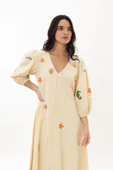 Vestido midi color arena con escote en V, mangas 3/4 abullonadas y cierre frontal con botones. Presenta bordados de motivos marinos.