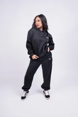 Conjunto deportivo negro con logo blanco en el pecho del hoodie y en el muslo del pantalón. El hoodie es de corte holgado con capucha y el pantalón es de corte recto con puños elásticos.