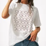 Camiseta blanca de algodón orgánico con cuello redondo y manga corta. Presenta un estampado frontal compuesto por círculos de puntos marrones, con letras pequeñas en el interior de algunos círculos.