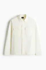 Sobrecamisa color crema con cuello inglés, cierre frontal con botones a presión, dos bolsillos superiores con solapa y puños ajustables con botón a presión.