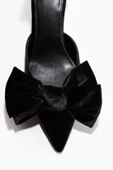 Zapatos de tacón tipo pump en terciopelo negro, con punta fina y un lazo grande decorativo en el empeine. Presentan una pulsera ajustable al tobillo con cierre de hebilla y tacón fino forrado.