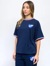 Remera blanca de corte clásico con cuello redondo y mangas cortas con puños tricolor (azul, blanco y rojo). Presenta el logo de Umbro bordado en el pecho izquierdo y el escudo del Club Nacional de Football bordado en el pecho derecho.