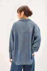Camisa de denim azul de corte amplio, manga larga, cuello camisero y bolsillos en el delantero. Presenta bordados dorados de mostacillas, lentejuelas y canutillos en mangas y parte inferior de un lateral del pantalón.