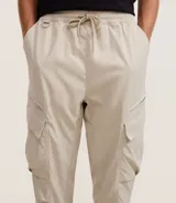 Pantalón cargo recto color beige, con cintura elástica ajustable con cordón, bolsillos laterales tipo cargo con solapa y bolsillos con cremallera.