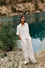 Camisa blanca de cuello solapa y manga corta, con bolsillo delantero y detalle de vainicas a tono. Cierre frontal con botones ocultos por solapa.