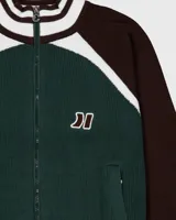 Campera de punto de algodón de grosor medio, con cierre frontal, cuello alto y puños y cintura en rib con franjas blancas y marrones. El cuerpo es verde oscuro y las mangas son marrones. Lleva un parche bordado con doble logo en el pecho.