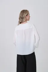 Blusa blanca de poliéster, entallada, con escote en V y botones frontales. Presenta mangas largas con puños anchos.