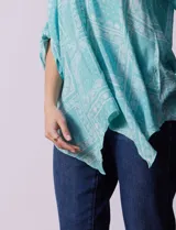 Blusa celeste con estampado de cachemir en blanco, cuello a la base, escote en V y abertura central con botones. Manga 3/4 regulable con presilla y botón, y terminación de ruedo asimétrico.